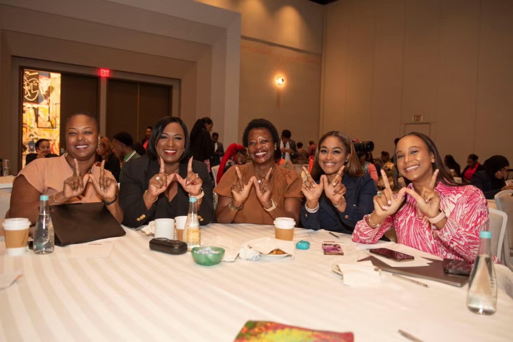 A group of five women sit around a table and smile to the camera, raising their fingers up in a "W"