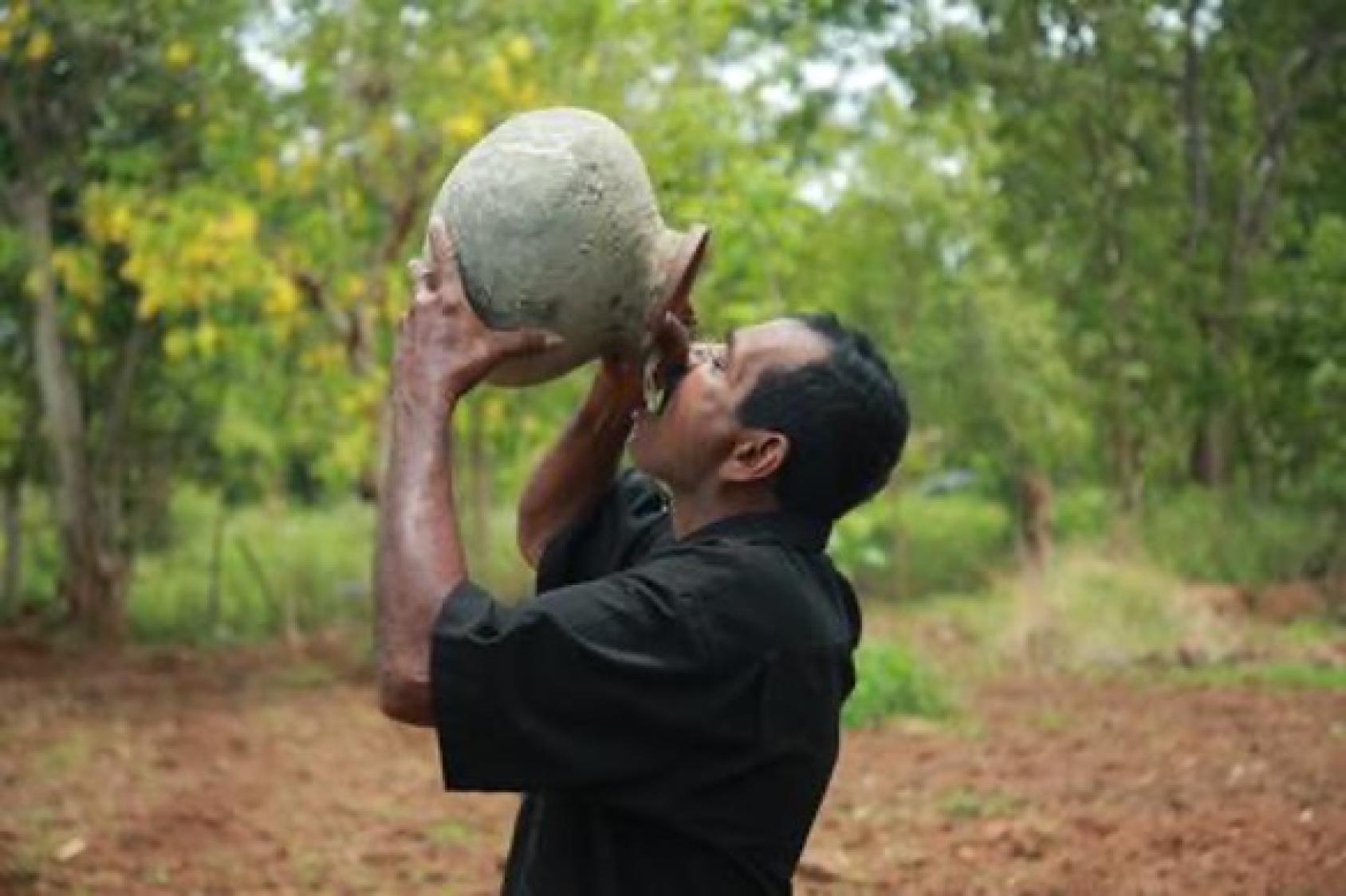 A man in a black shirt tips over an earthen pot filled with water in his mouth.
