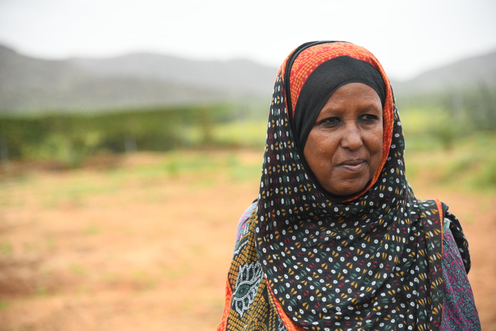 A woman in a black and orange printed headscarf against a landscape with mountains in the distance.