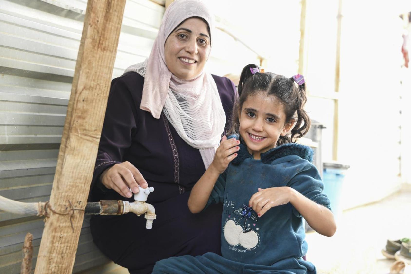 A small girl in a blue dress and an older woman in a black dress and a pink headscarf sit next to a water pipe with a tap.
