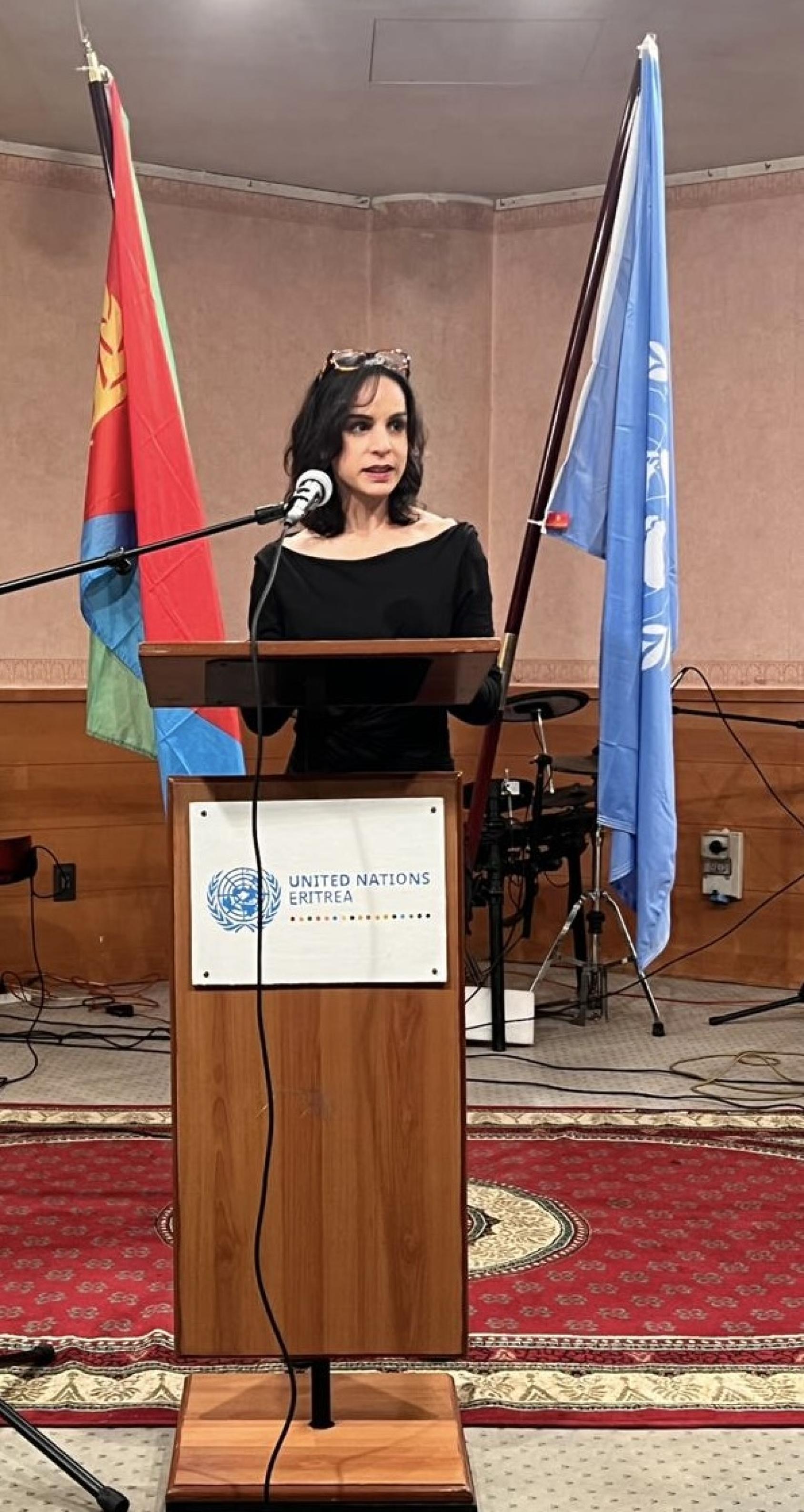 A woman with dark hair, wearing a black shirt speaks into a microphone at a wooden podium. Behind her are two flag, one of the country of Eritrea and another a blue UN flag