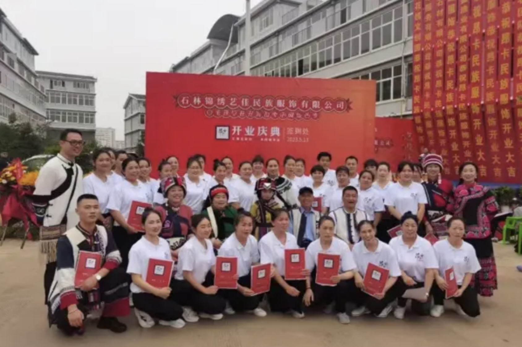 一群人在一个红色的标牌前方合影，第一排蹲着第二三排站着