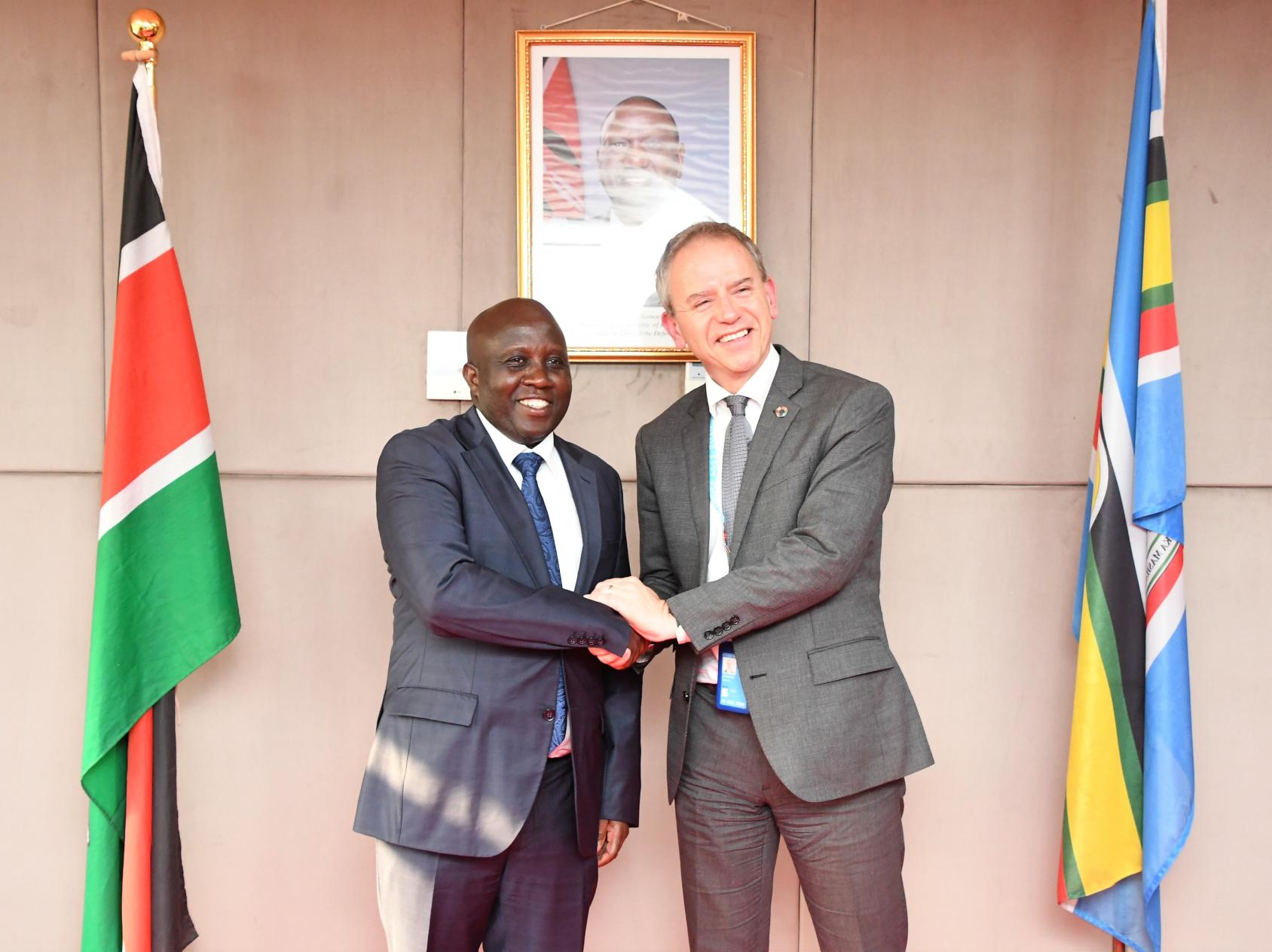 Two men in grey suits shake hands as they stand in front of a panelled wall with the flag of Kenya on it