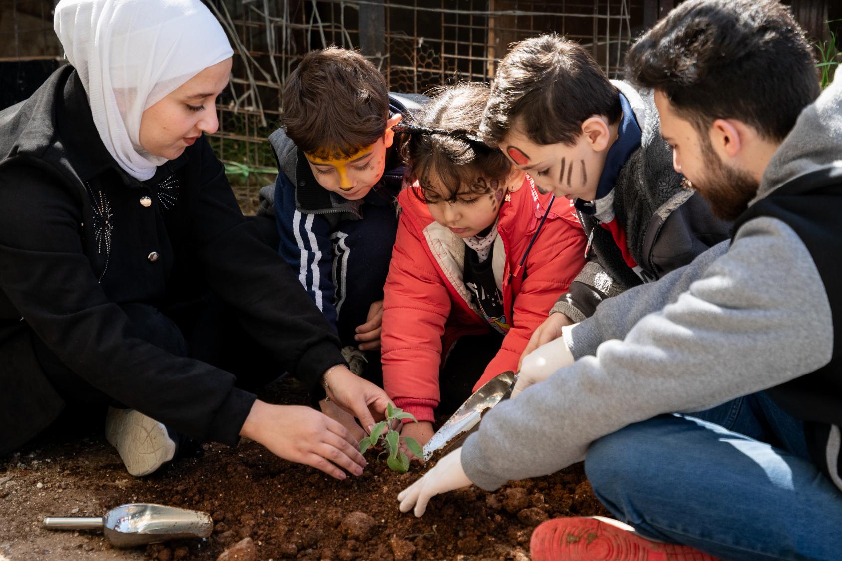 A group of three children and two adults plant a sapling into the ground.