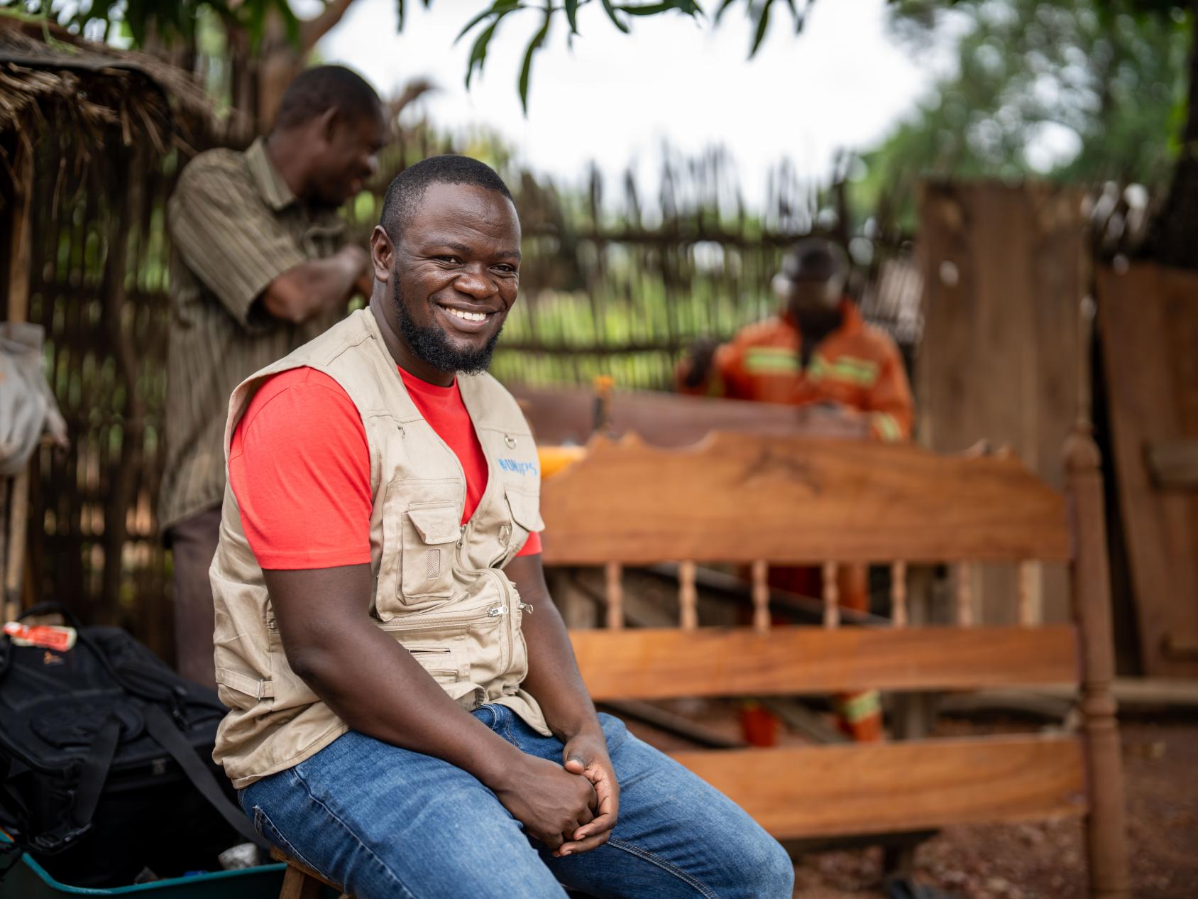 A man in a red shirt and a grey vest sits atop a wooden bench