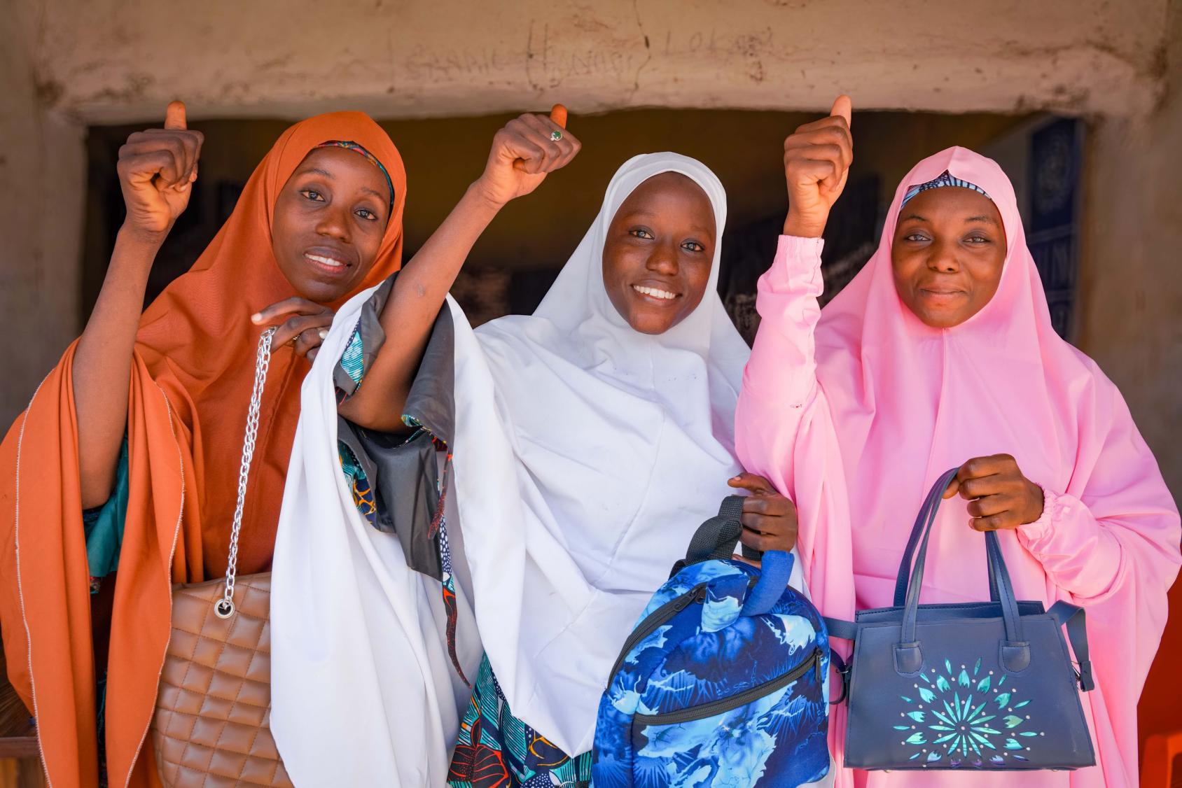Three women in orange, white and pink clothes stand side by side with their fingers raised in a thumbs-up sign.