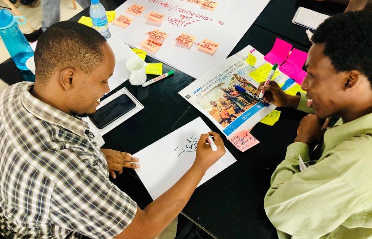 Deux jeunes hommes africains sont assis à un bureau de couleur noire couvert de post-its, de feuilles de papier et d'objets divers. Ils réfléchissent ensemble et esquissent des solutions sur une feuille de papier en souriant.
