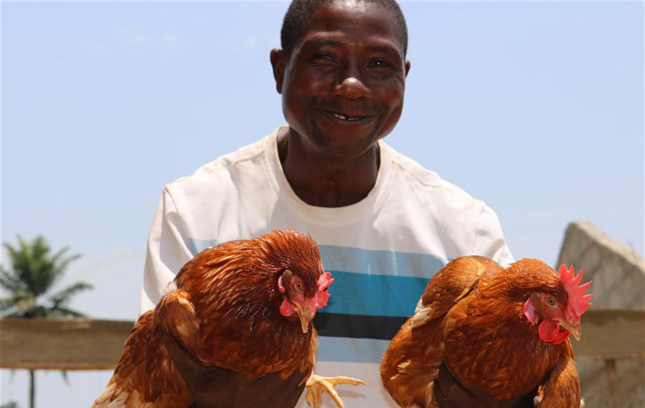 Un homme sourit à la caméra en tenant deux coqs dans les mains