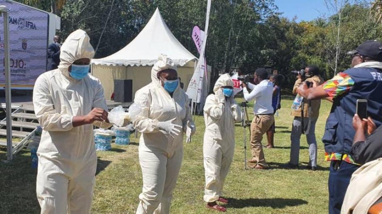 Plusieurs caméramans et photographes se tiennent devant une rangée de personnes distantes les unes des autres et vêtues de combinaisons de protection blanches, de gants, de charlottes et de masques chirurgicaux bleus.
