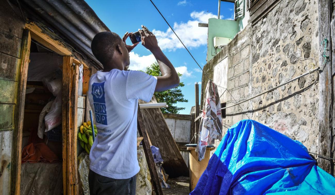 Dans une ruelle exigüe jonchée d’objets en tous genres, un jeune homme vêtu d’un t-shirt au dos duquel on peut voir le logo du PNUD prend en photo le mur qui se trouve en face de lui à l’aide de son téléphone mobile.