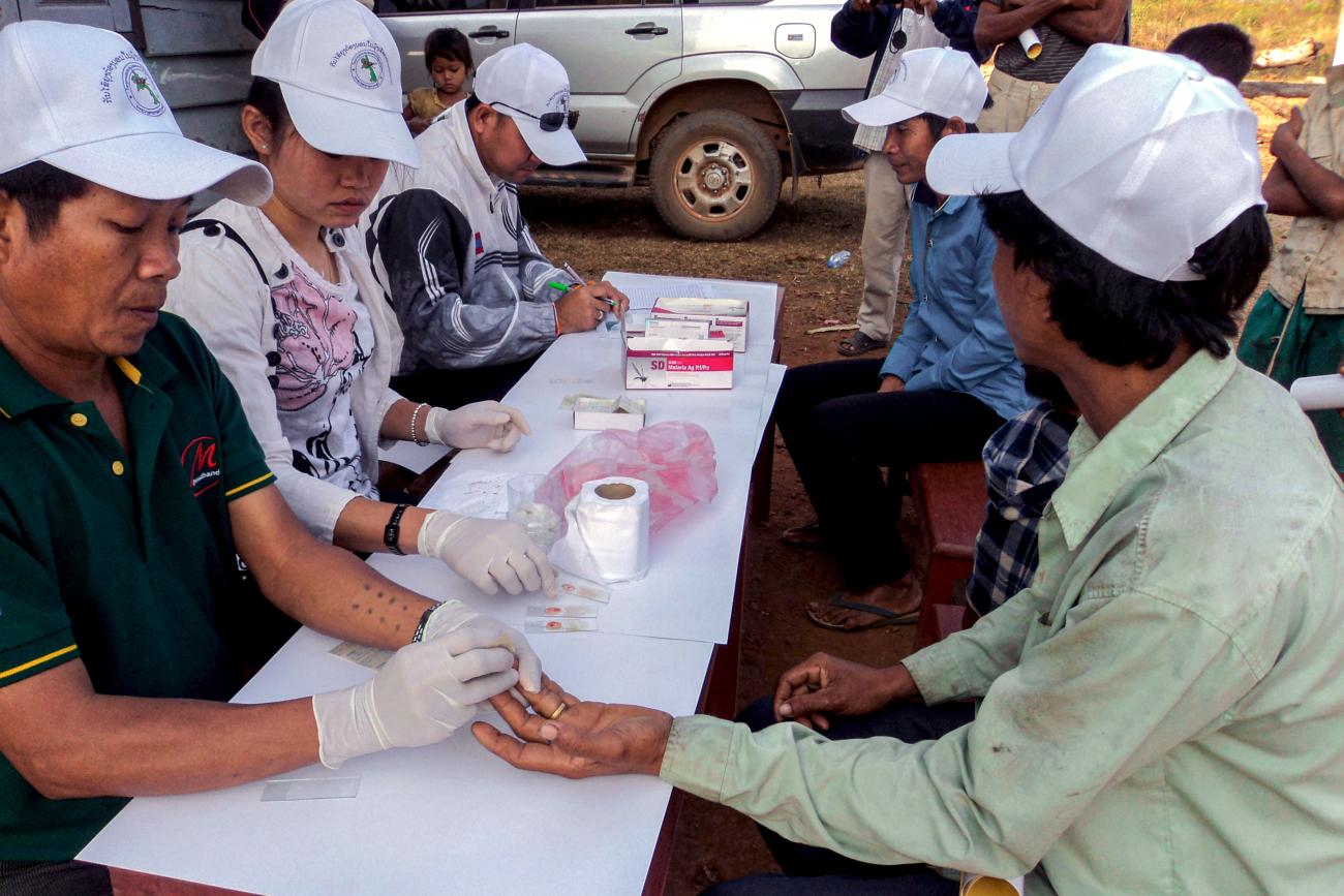 Plusieurs personnes portant des casquettes blanches sont assises en extérieur à une table sur laquelle sont posés des kits de dépistage du paludisme. En premier plan, un agent de santé réalise un test de diagnostic rapide du paludisme chez l’homme assis en face de lui en lui piquant l’annulaire gauche pour obtenir une goutte de sang.