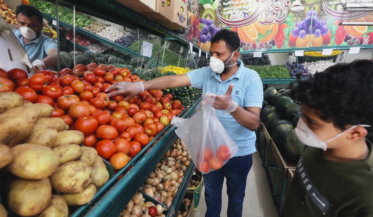 Abdou, un réfugié yéménite à Amman, fait ses courses chez un marchand de légumes après la réouverture des petits commerces. Il porte un masque de protection et des gants. Au premier plan de l’image, un enfant, lui aussi portant un masque, le regarde mettre des tomates un sac en plastique.