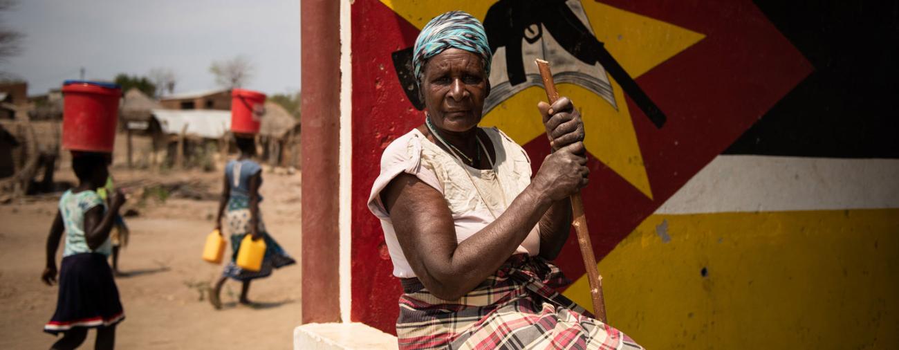 Una mujer, que huyó del conflicto, se sienta frente a un mural. Ella sostiene un palo y mira estoicamente hacia la cámara. A la izquierda de la imagen, al fondo, dos niñas caminan una detrás de la otra, cada una con un balde rojo en la cabeza.