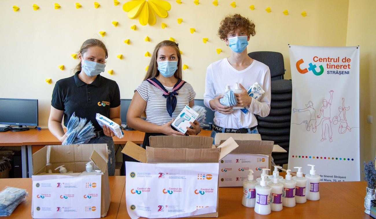 Trois jeunes bénévoles, deux jeunes filles et un jeune homme, remplissent des cartons contenant de produits de première nécessité destinés à être distribués. Ils se tiennent derrière un comptoir sur lequel sont posés des cartons et divers produits. Chacun porte un masque de protection.