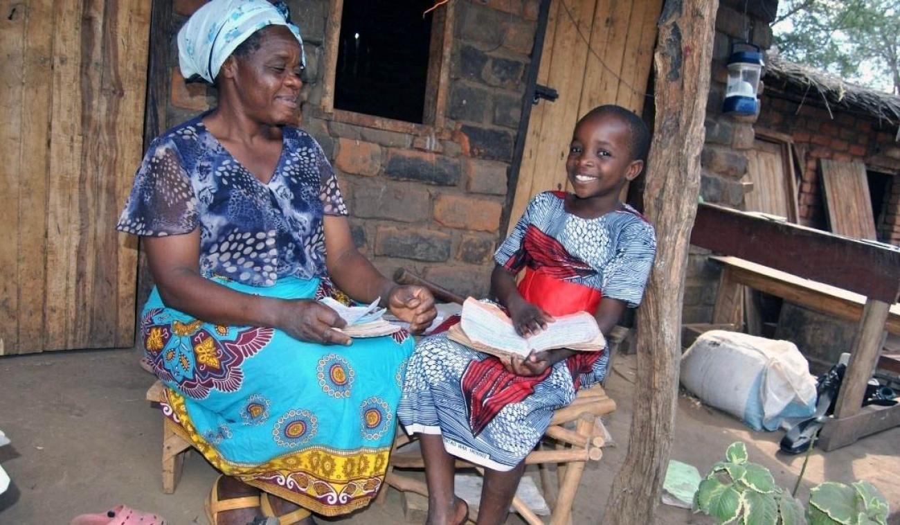 Une mère aide sa fille à faire ses devoirs. Elles sont assises côte à côte sur des chaises en bois devant ce qui semble être une cabane faite de pierre et de bois. La fille, à droite, a le dos appuyé contre un poteau en bois. Elle tient un cahier dans les mains et regarde la caméra avec un grand sourire. Sa mère, à gauche, tient des feuilles de papier et regarde dans la direction de sa fille en souriant discrètement.