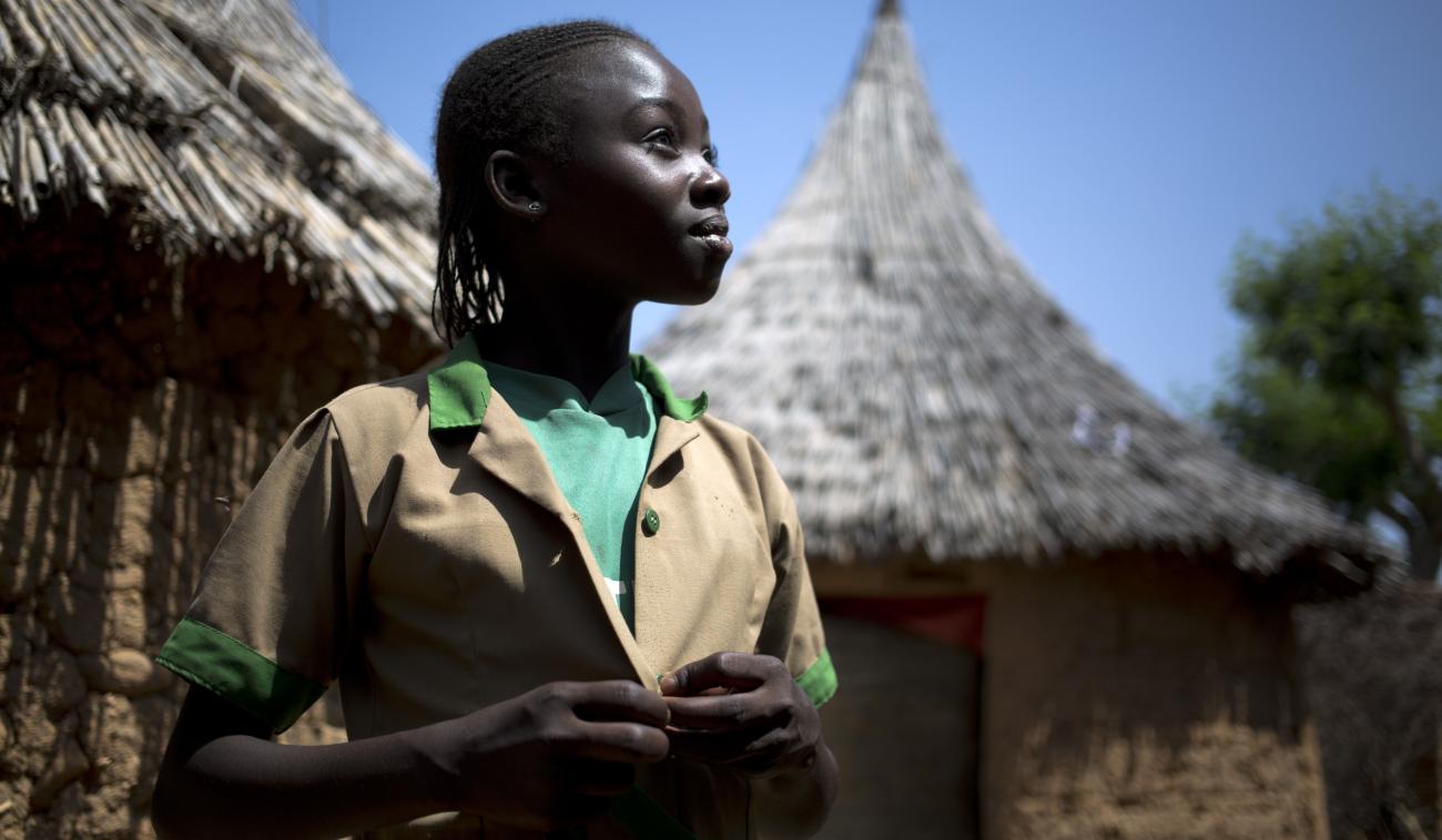Une jeune fille se tient debout en regardant droit devant elle d’un air fier. Derrière elle, on perçoit deux huttes.