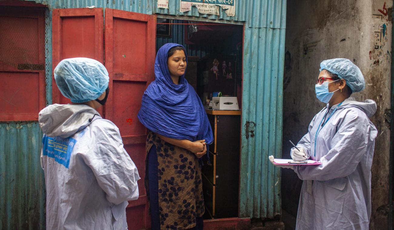 Trois femmes se tiennent devant la devanture bleue et rouge d’un magasin traditionnel. L’une d’elle est une riveraine. Elle porte une robe et une grande étole bleue autour de la tête et des épaules. Les deux autres femmes sont des travailleuses sociales. Elles portent chacune un masque et un équipement de protection individuelle. L’une des deux travailleuses sociales prend des notes dans un carnet.