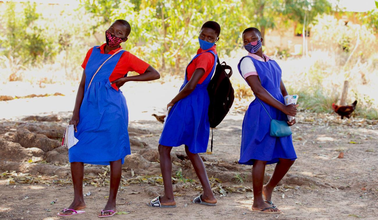 Trois écolières portant un masque de protection s'amusent dans la cour de l'école primaire de Luwambaza, en prenant la pose pour la caméra. Toutes trois ont les cheveux courts et portent l’uniforme de l’école, une tenue bleu vif.