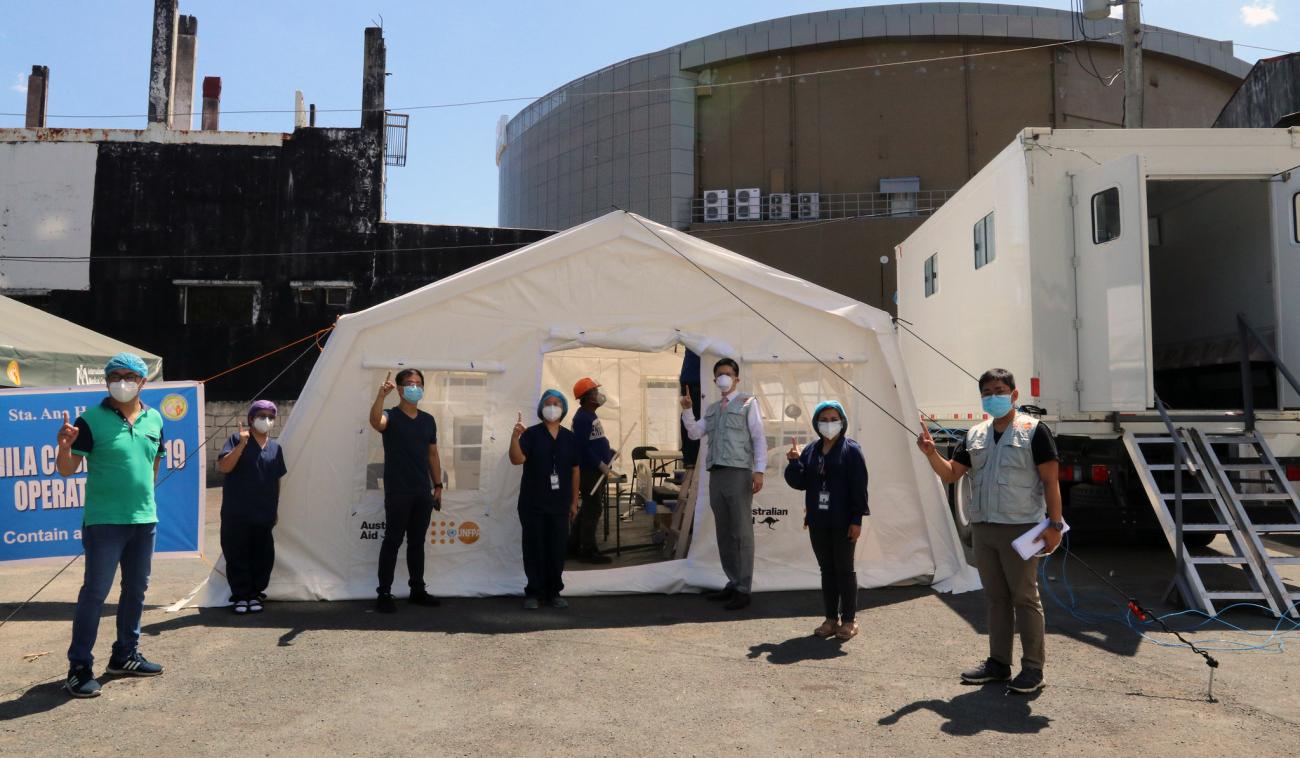 Des membres du personnel de l'ONU, des fonctionnaires locaux et des représentants de partenaires locaux se tiennent debout, face à la caméra, à l'extérieur d’une grande tente blanche offerte par le FNUAP pour le triage des patients COVID-19 à l'hôpital Sta. Ana, aux Philippines. Tous portent un équipement de protection individuelle et un masque, respectent les distances de sécurités recommandées et pointent l’index vers le haut.