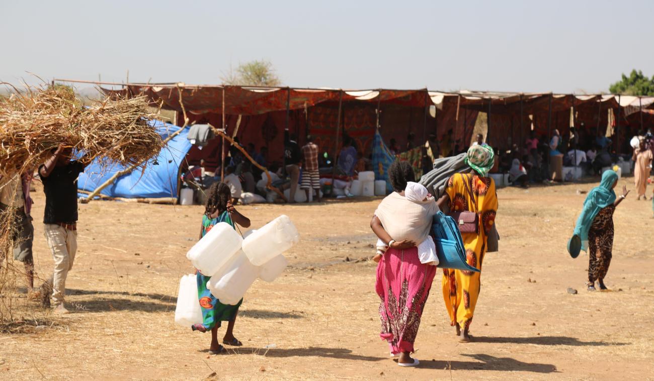 Dos mujeres y una niña caminan hacia un campamento, de espaldas a la cámara, cargando latas, colchones y mantas. Al fondo, se puede ver a muchas personas refugiadas en tiendas de campaña.