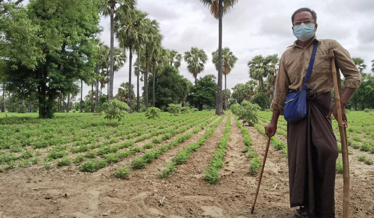 Un agricultor, a la derecha de la imagen, está de pie junto a un campo agrícola verde. Lleva una máscara protectora y se apoya en muletas.