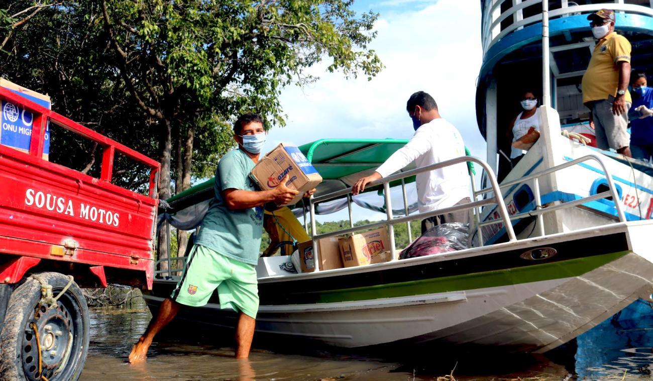 Un hombre en pantalones cortos y con una máscara protectora descarga los suministros entregados por la OIM en un litoral. Se le ve moviéndose, cargando una caja de cartón, entre una camioneta roja y un bote pequeño. Detrás de la primera embarcación, podemos ver otro barco más grande con varias personas a bordo. Todos los presentes usan mascarillas.