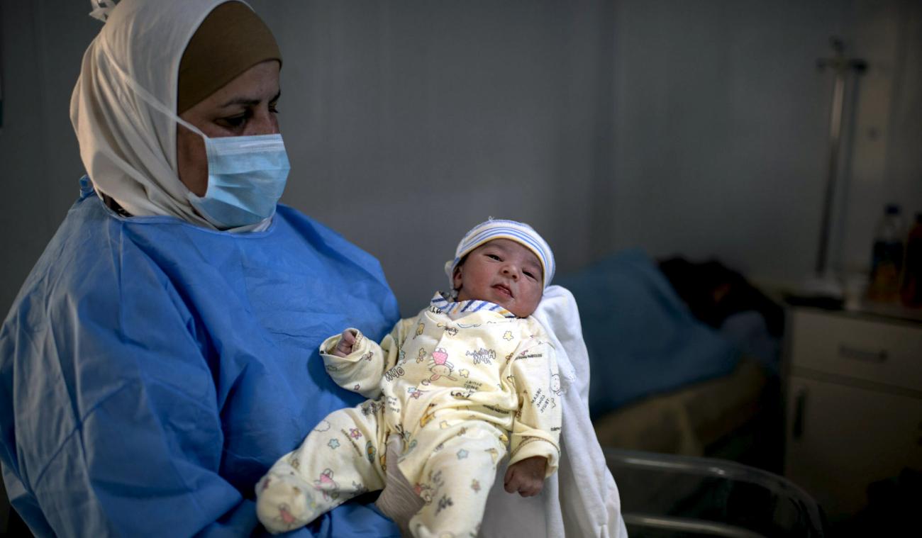 Une soignante portant un masque et une blouse de protection bleue tient un nouveau-né dans les bras.
