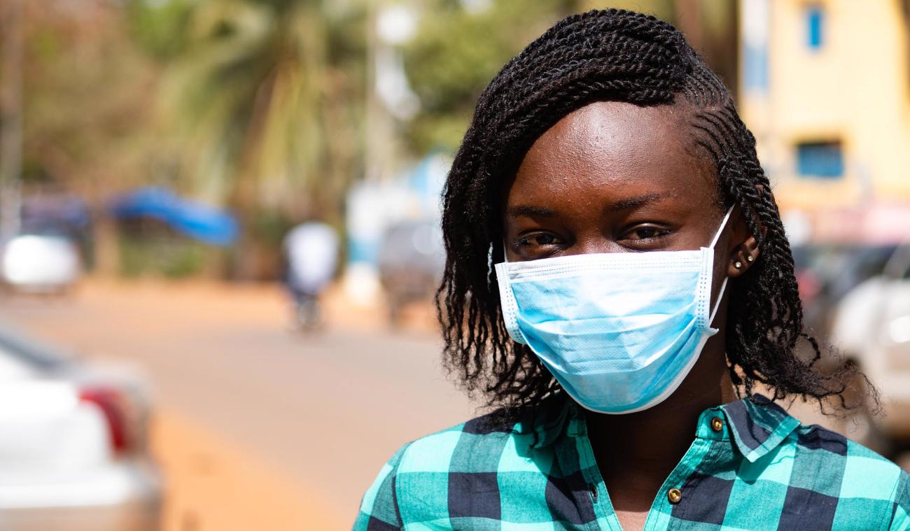 A mid-close up of an adolescent girl wearing a face mask poignantly looking at the camera. 