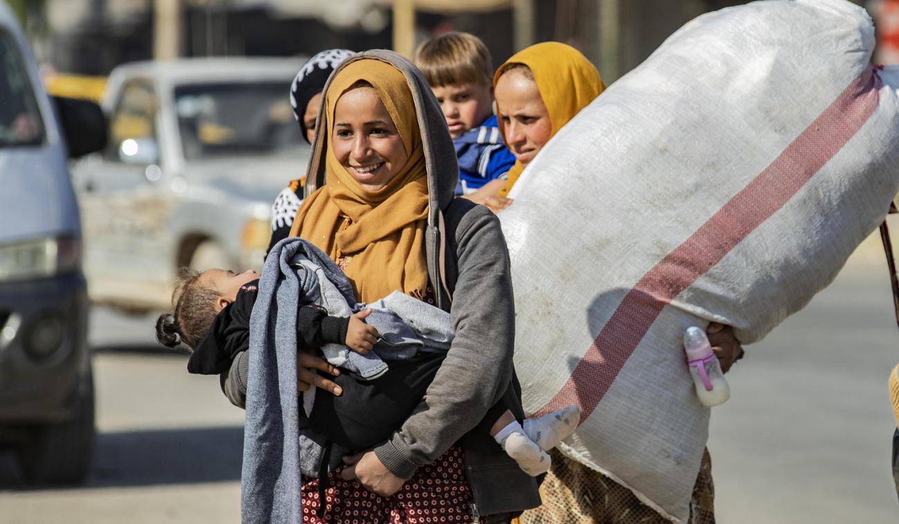 Une femme sourit tandis qu'elle porte un enfant endormi dans les bras. Une autre femme portant un très grand sac marche derrière elle.