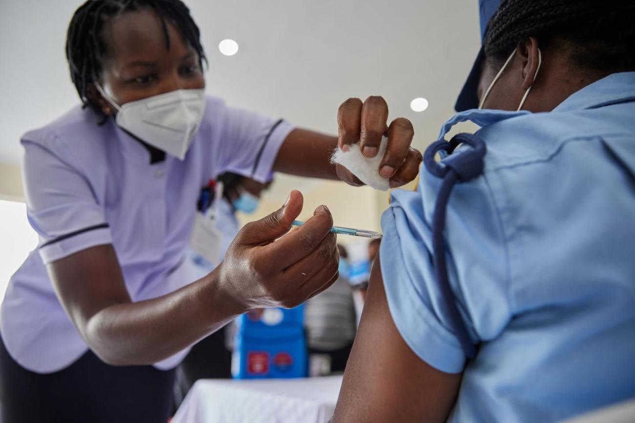 A nurse in a purple shirt and white face mask administers a covid vaccine on a woman with a blue shirt and cap.