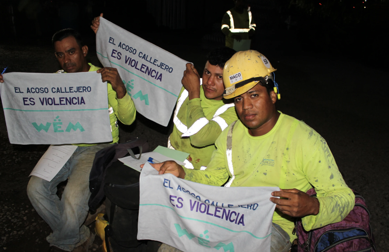 Trois hommes portant des tenues de travail jaunes regardent la caméra en tenant chacun entre les mains une serviette sur laquelle on peut lire, en espagnol : " El acoso calejero es violencia", ce qui signifie en français : "Le harcèlement de rue est une violence ".
