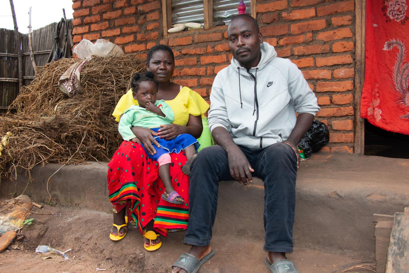 Un homme et une femme portant une fillette sur les genoux sont assis sur une marche devant une maison en briques.
