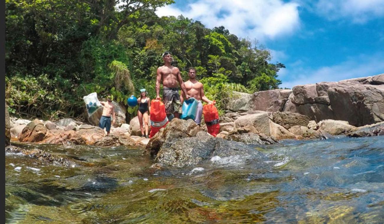 Quatre personnes en maillot de bain et tenant du matériel dans les mains se tiennent debout sur des rochers sur une côte brésilienne. On aperçoit derrière eux une végétation luxuriante.