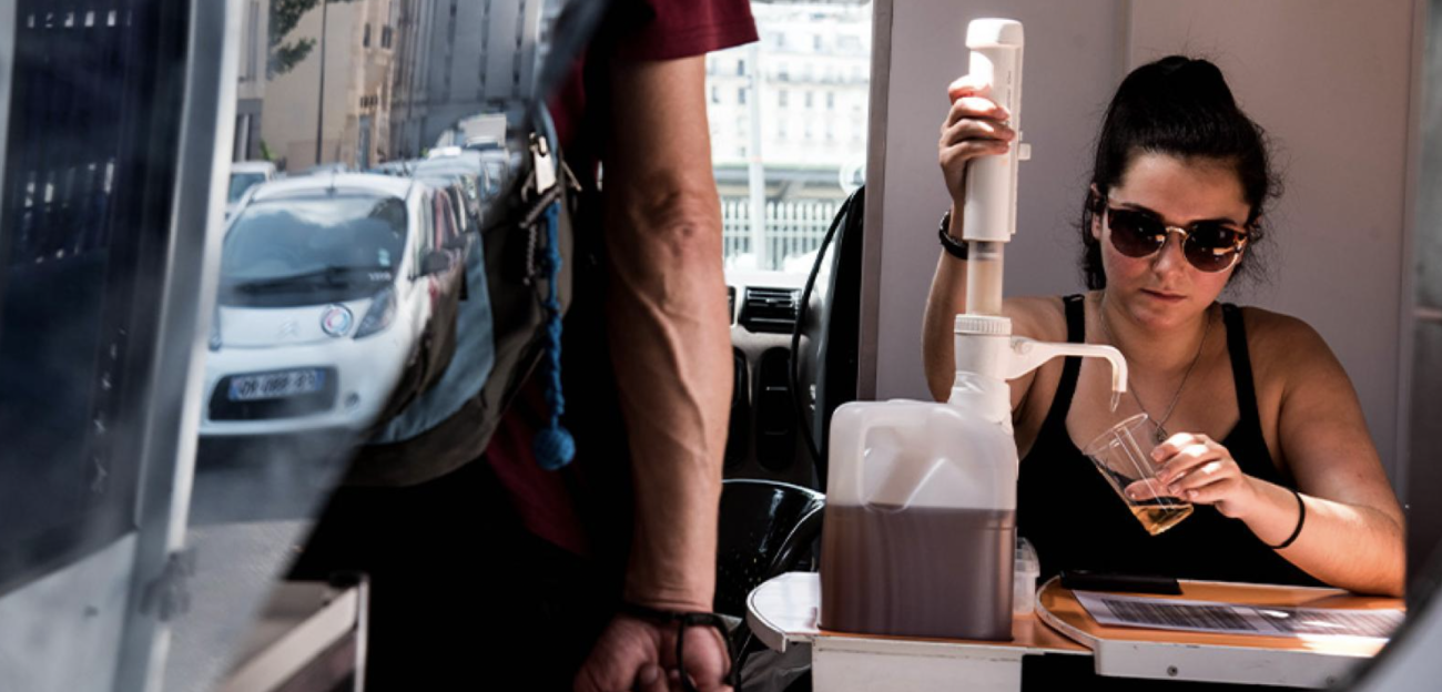 Une femme portant des lunettes de soleil est assise à une table et verse un liquide brunâtre dans une tasse à partir d'un récipient en plastique.