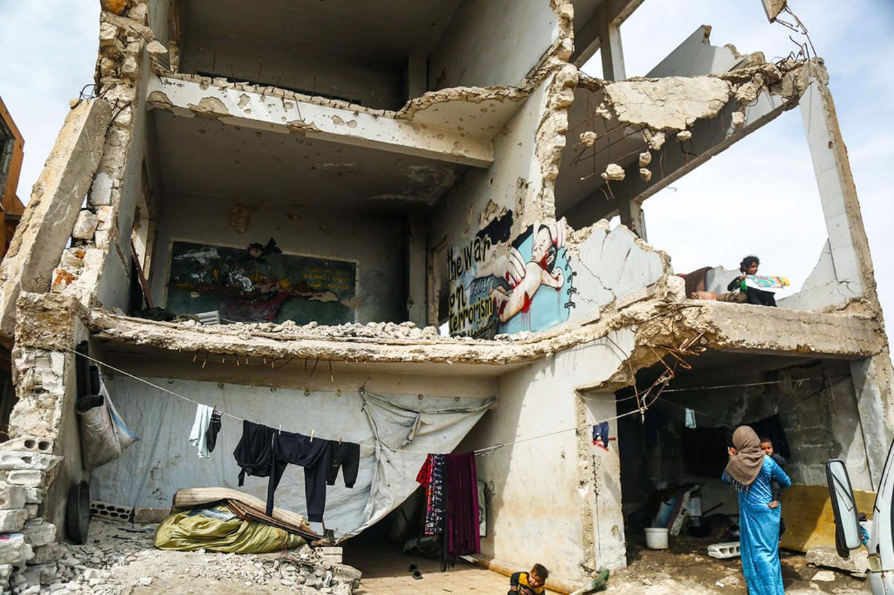 Une femme vêtue d’une robe bleue et portant un enfant dans les bras se tient devant un bâtiment dont la façade a été complètement détruite. Un autre enfant est accroupi près d’elle et une fillette se tient près d’un rebord, au premier étage du bâtiment.