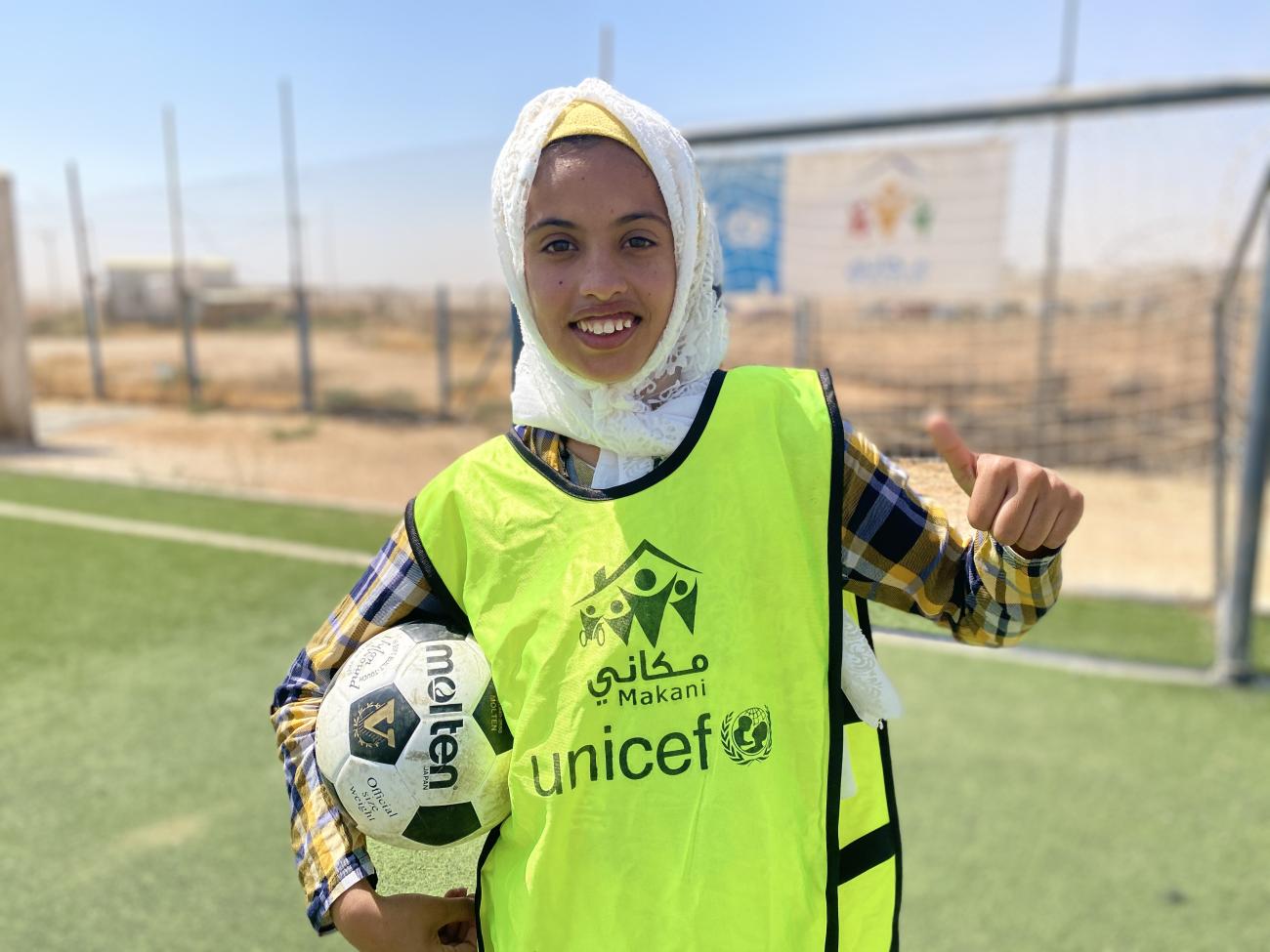 A smiling young girl holds a soccer ball in one arm and gives the camera a thumbs up with her other hand. 
