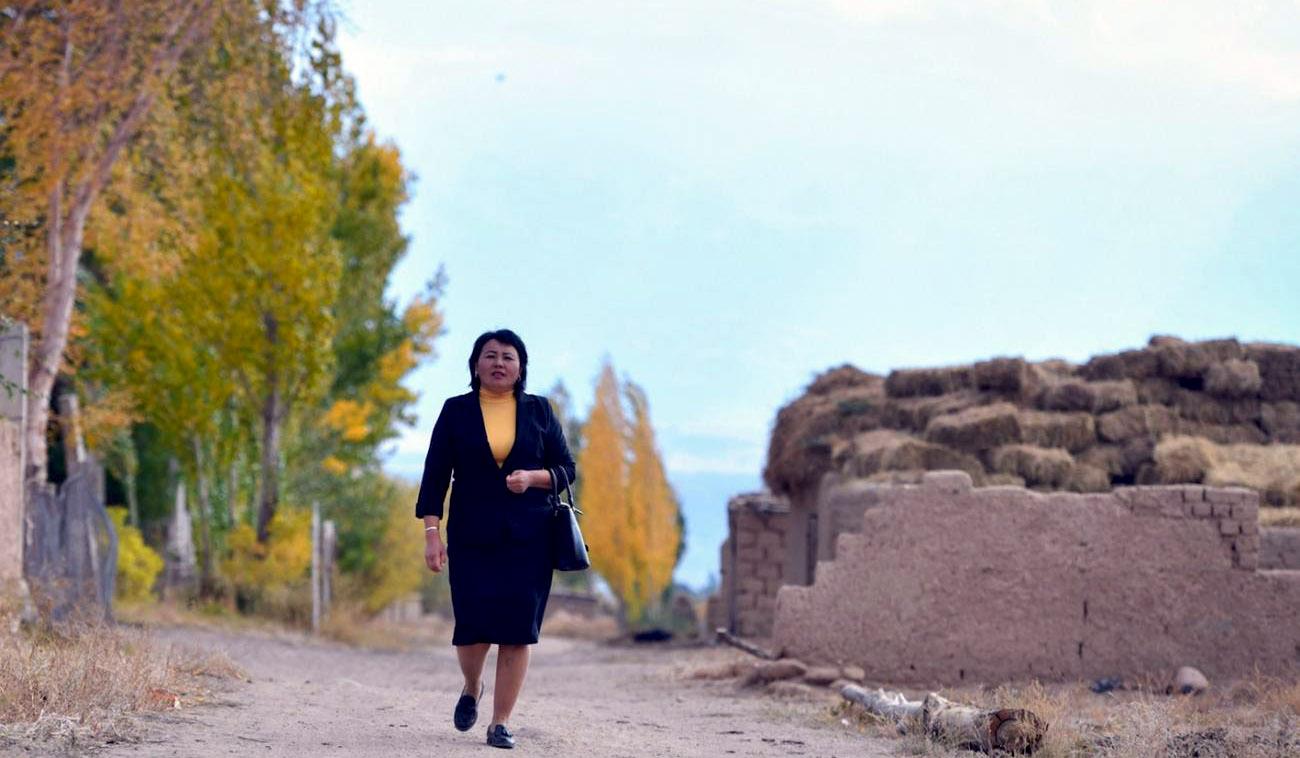 Une femme en tailleur noir marche sur une route de village en direction de la caméra.