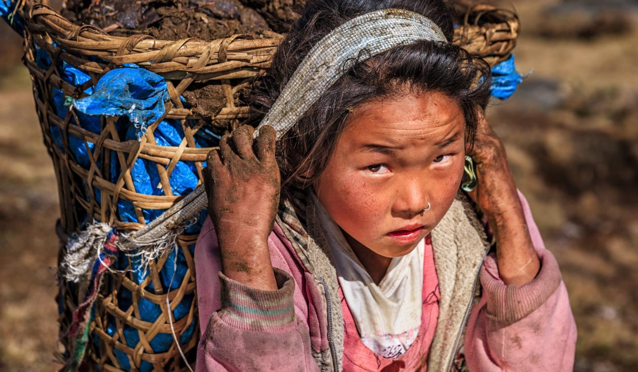 A young girl, with dirt on her hands and face, carries a large basket on her back.
