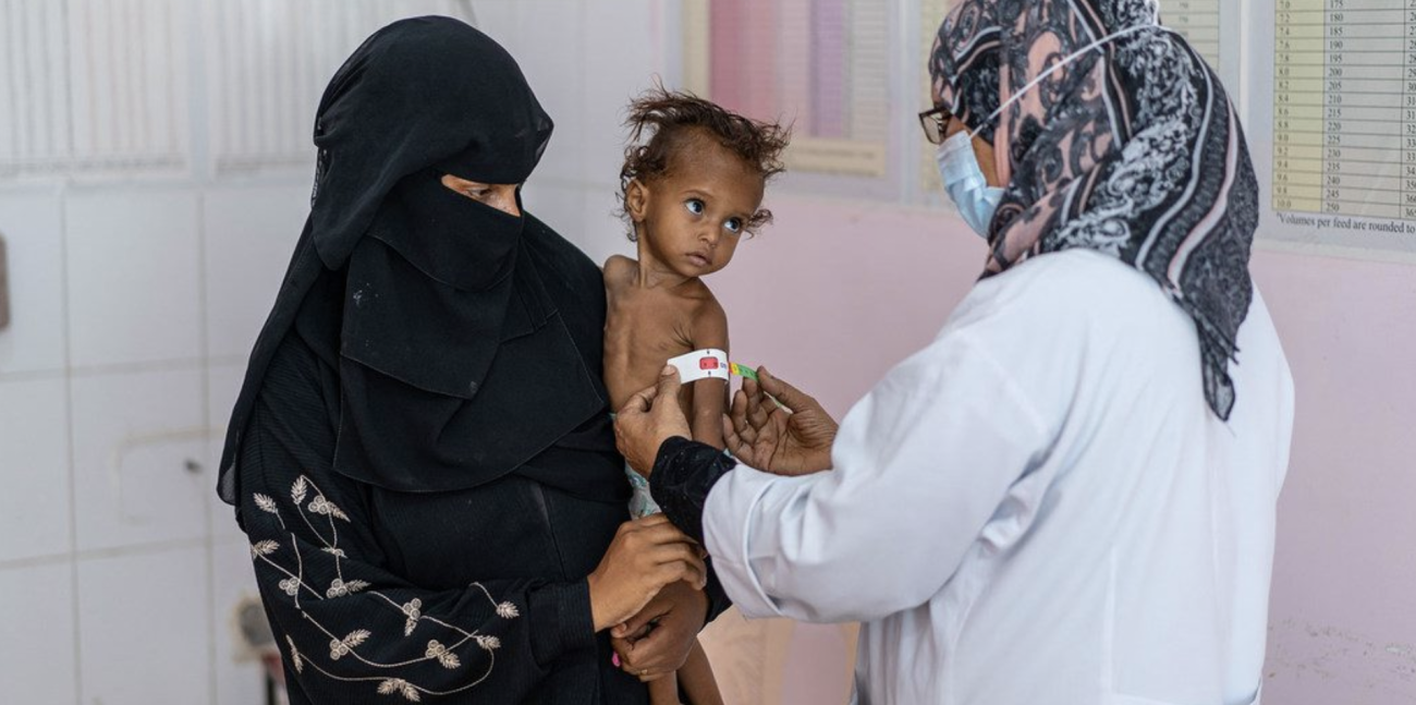 Une femme portant un voile noir lui couvrant le visage tient un enfant atteint de malnutrition sévère dans les bras, tandis qu'une infirmière mesure le diamètre du bras gauche de l'enfant.