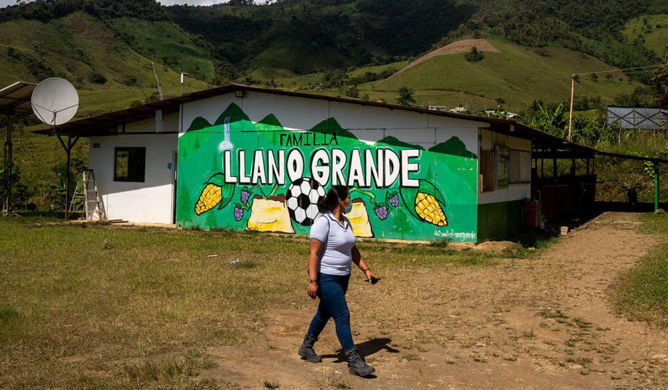 A woman wearing a face mask walks in front of a house with a painted green mural of food and a soccer ball. 