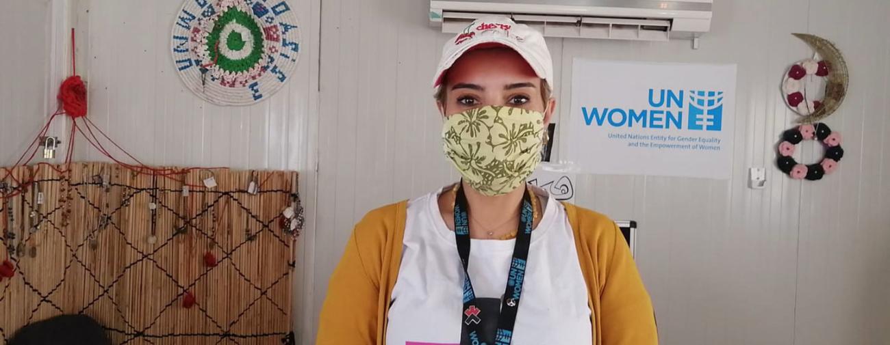 امرأة ترتدي كمامة تنظر مباشرة إلى الكاميرا بجوار لافتة هيئة الأمم المتحدة للمرأة.