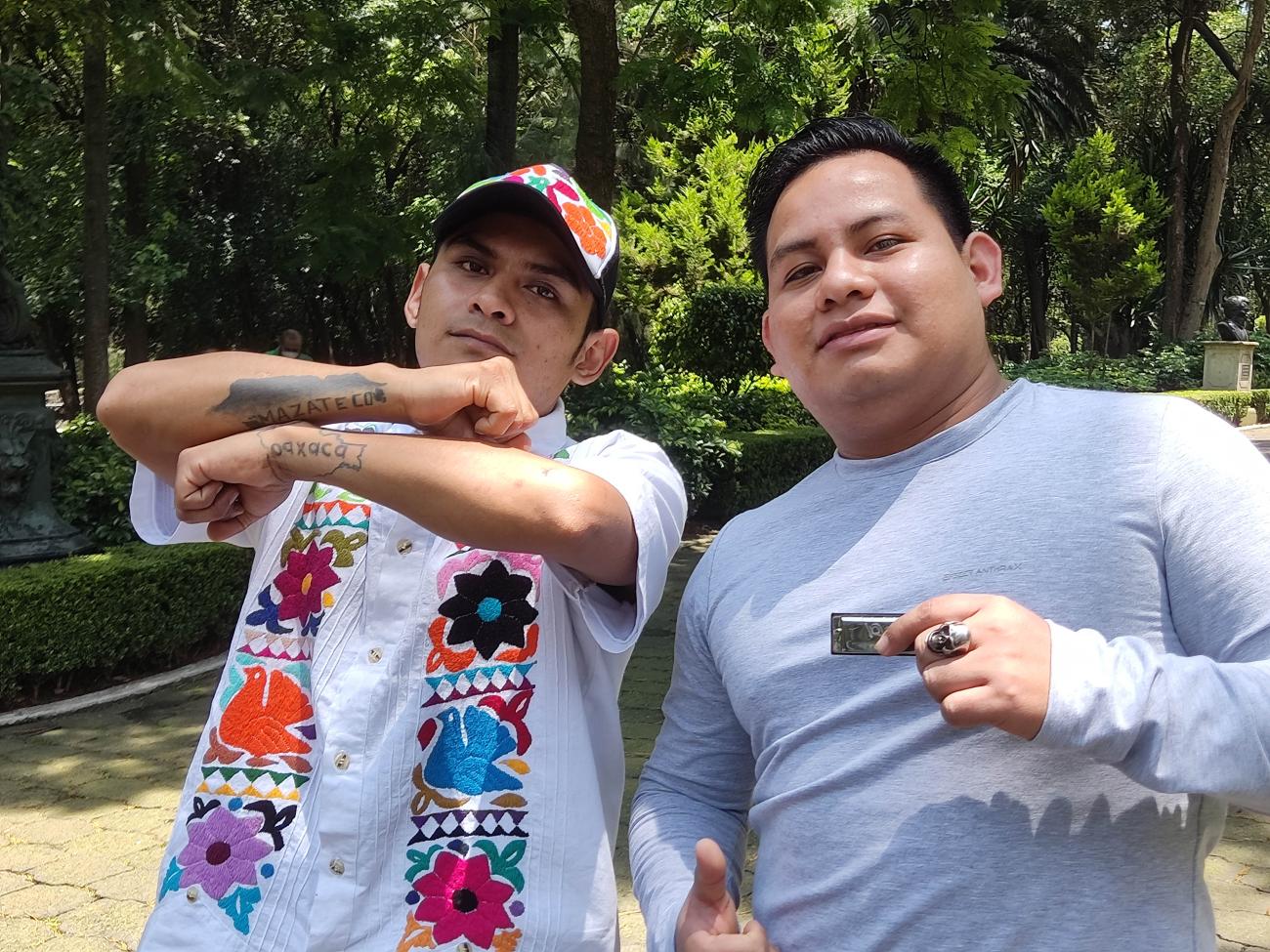 Two men stand together near a wooded area. 