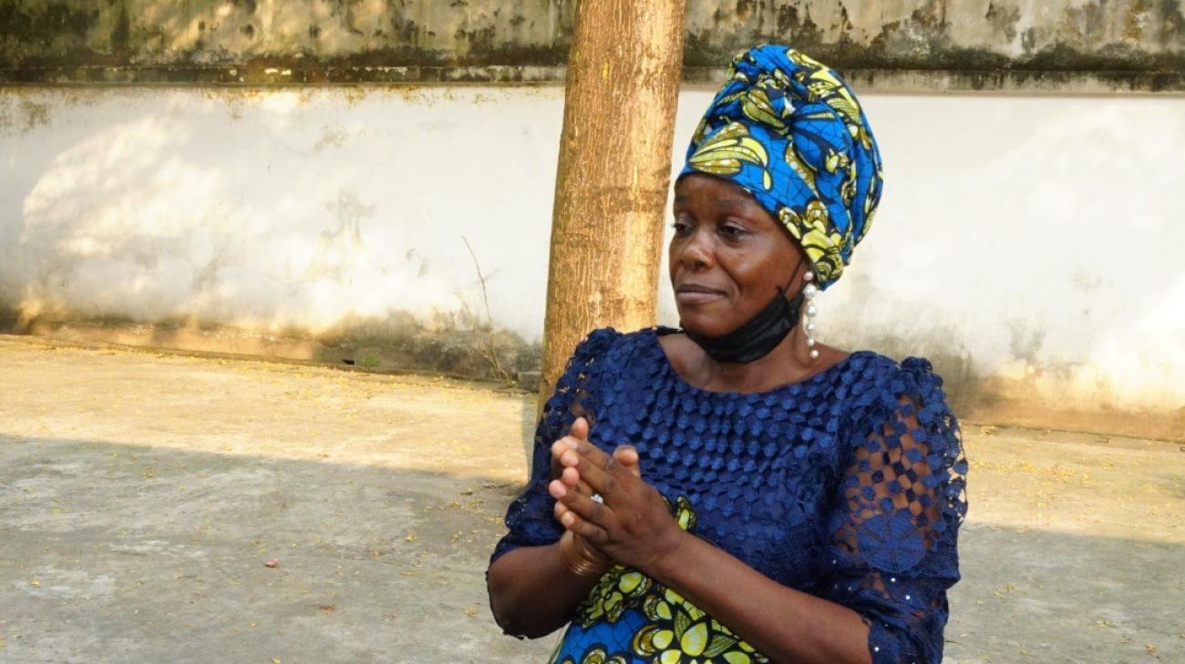 A woman in a blue dress stands with her hands together on a street. 