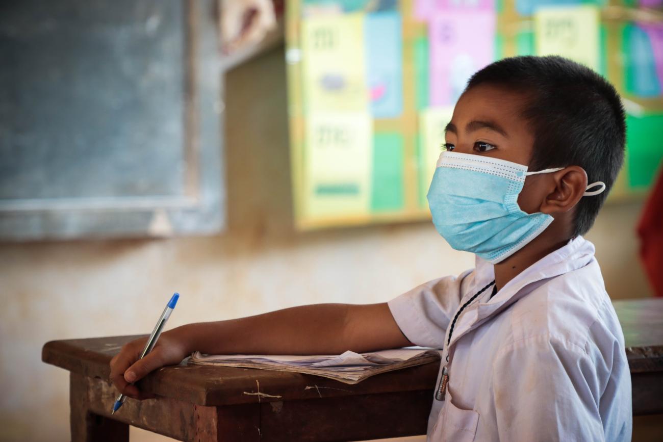 Un joven estudiante, usando mascarilla en un salón de clases, mira hacia la lado, mientras toma apuntes con un bolígrafo.