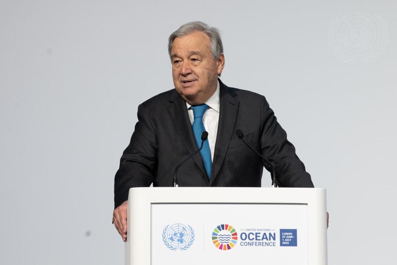 A man in a suit speaks into a microphone and a podium with UN logos on it, against a grey wall. 