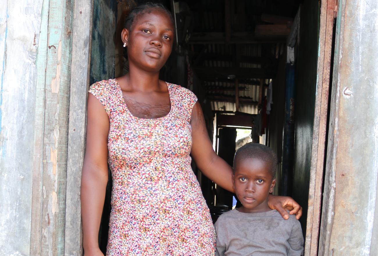 امرأة شابة تقف على عتبة بابها وذراعها حول كتف ابنها الصغير.