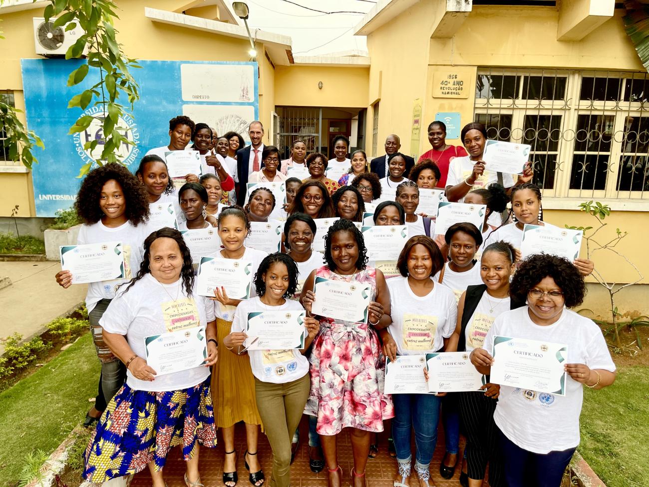 Mujeres con mangas de camisa blanca sostienen certificados en sus manos, frente a un edificio municipal, con el Coordinador Residente de pie en el centro-fondo.