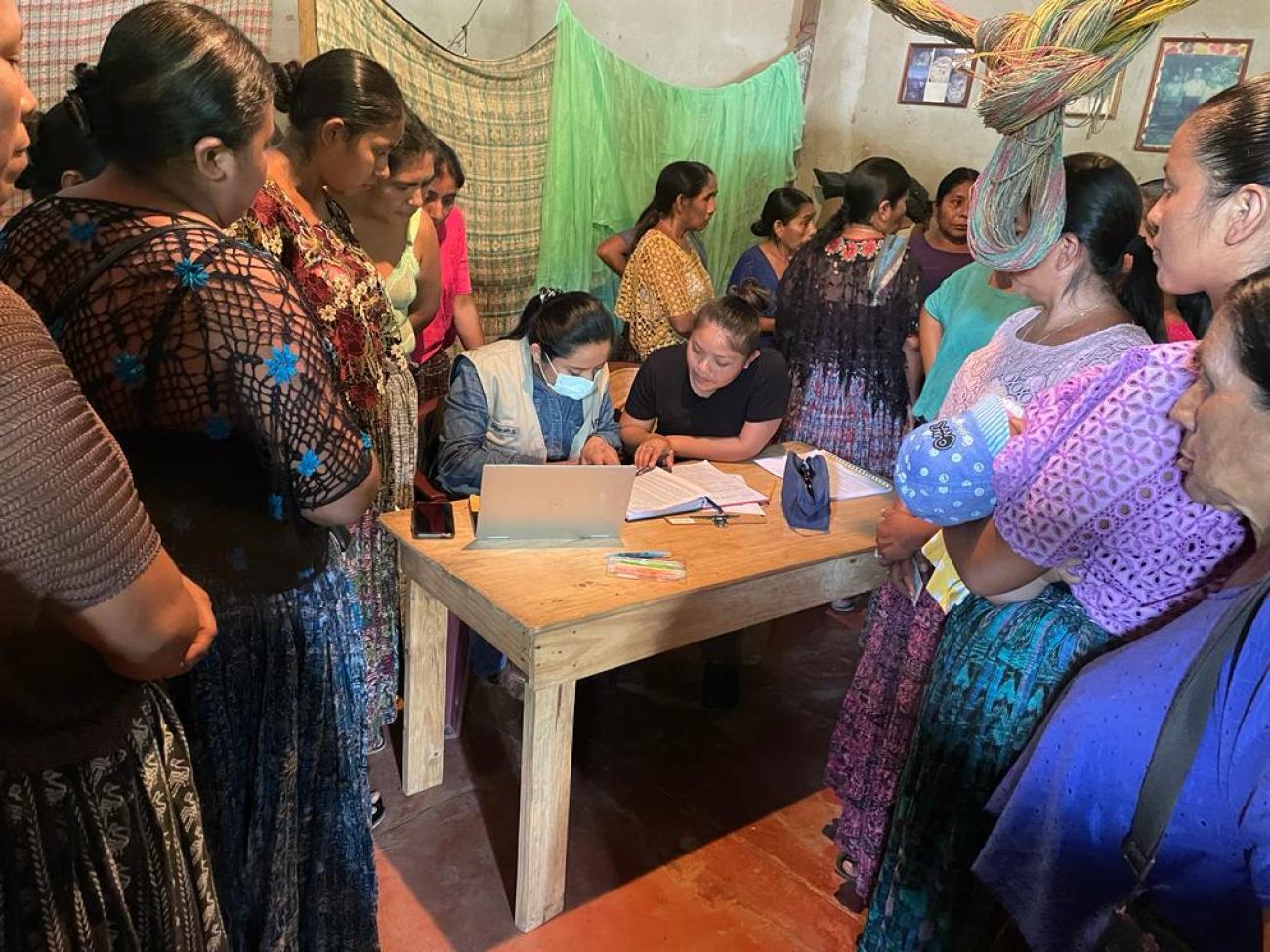 A group of women gather around two women who are looking at different notebooks and the screen of the laptop that lays on a wooden table.