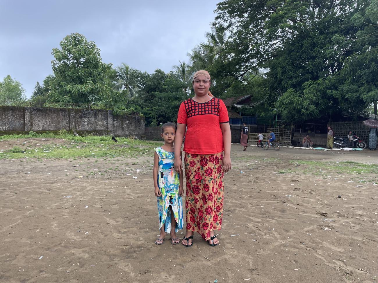 A woman stands next to a young girl, on a dry patch of land with some grass, trees, and people on motorbikes behind them.