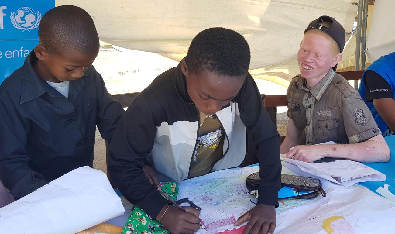 En Madagascar, dos niños estudian en una mesa bajo una gran tienda de campaña instalada por UNICEF, mientras un tercer niño, albino, les observa con una gran sonrisa.